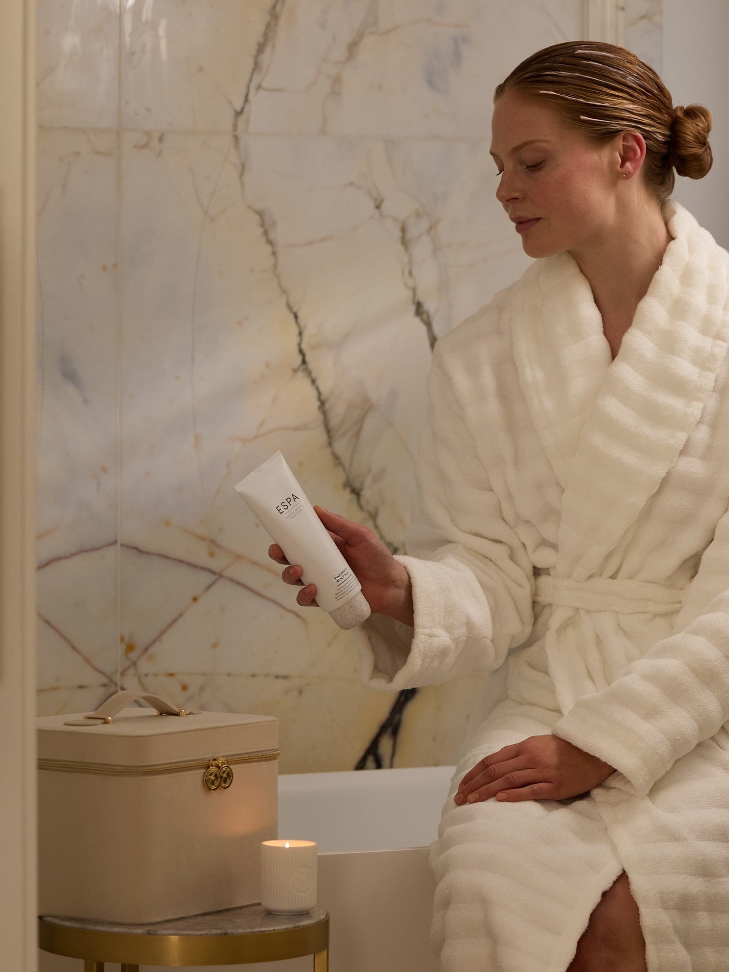Image of woman sat on side of bath with a white robe on. Hair in a hair mask with the cream vanity case on a side table with a small candle lit