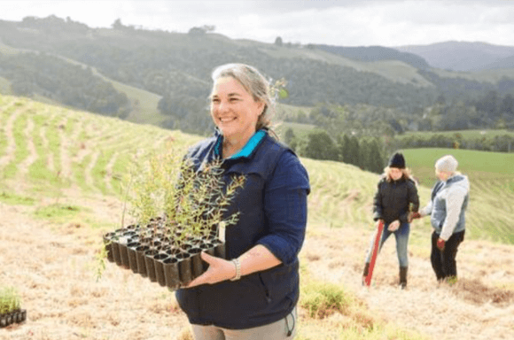 Woman holding plants that are going to planted to offset carbon with two ladies behind her digging up the ground.